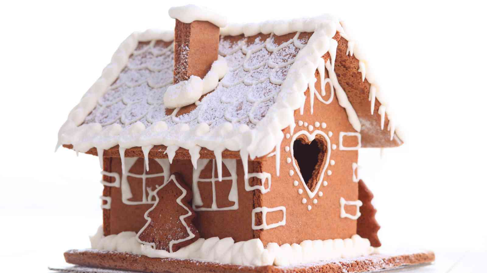 A gingerbread house decorated with white icing, featuring a heart-shaped window, a tree, and snow-like details on the roof and edges.