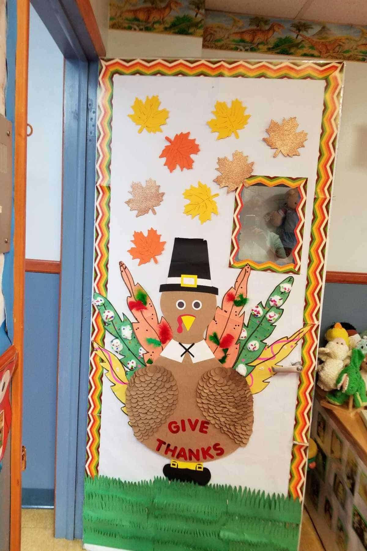 Classroom door decorated with a large paper turkey, colorful feathers, leaves, and the words "GIVE THANKS," celebrating Thanksgiving.