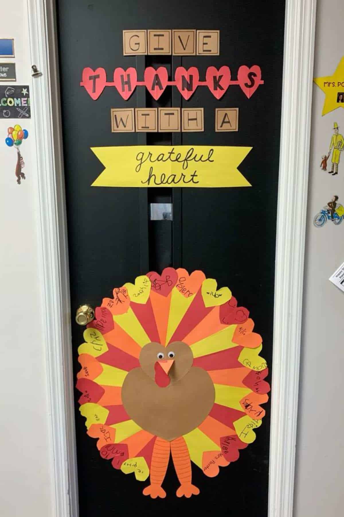 A black door decorated with a paper turkey and the words "Give Thanks With A Grateful Heart." The turkey's feathers have handwritten notes listing things to be thankful for.