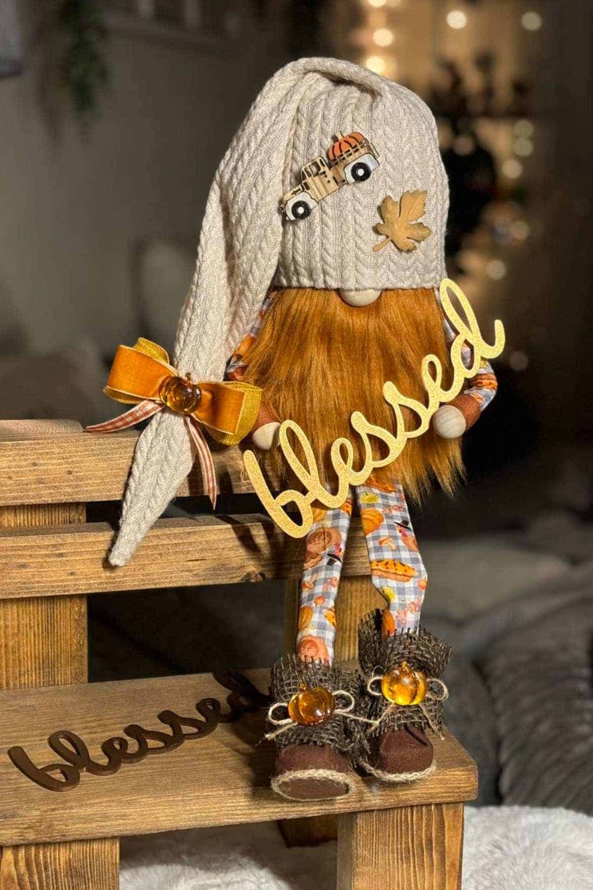 A gnome doll with a long hat, orange beard, and autumn-themed clothes holds a "blessed" sign while sitting on a wooden bench with another "blessed" sign.