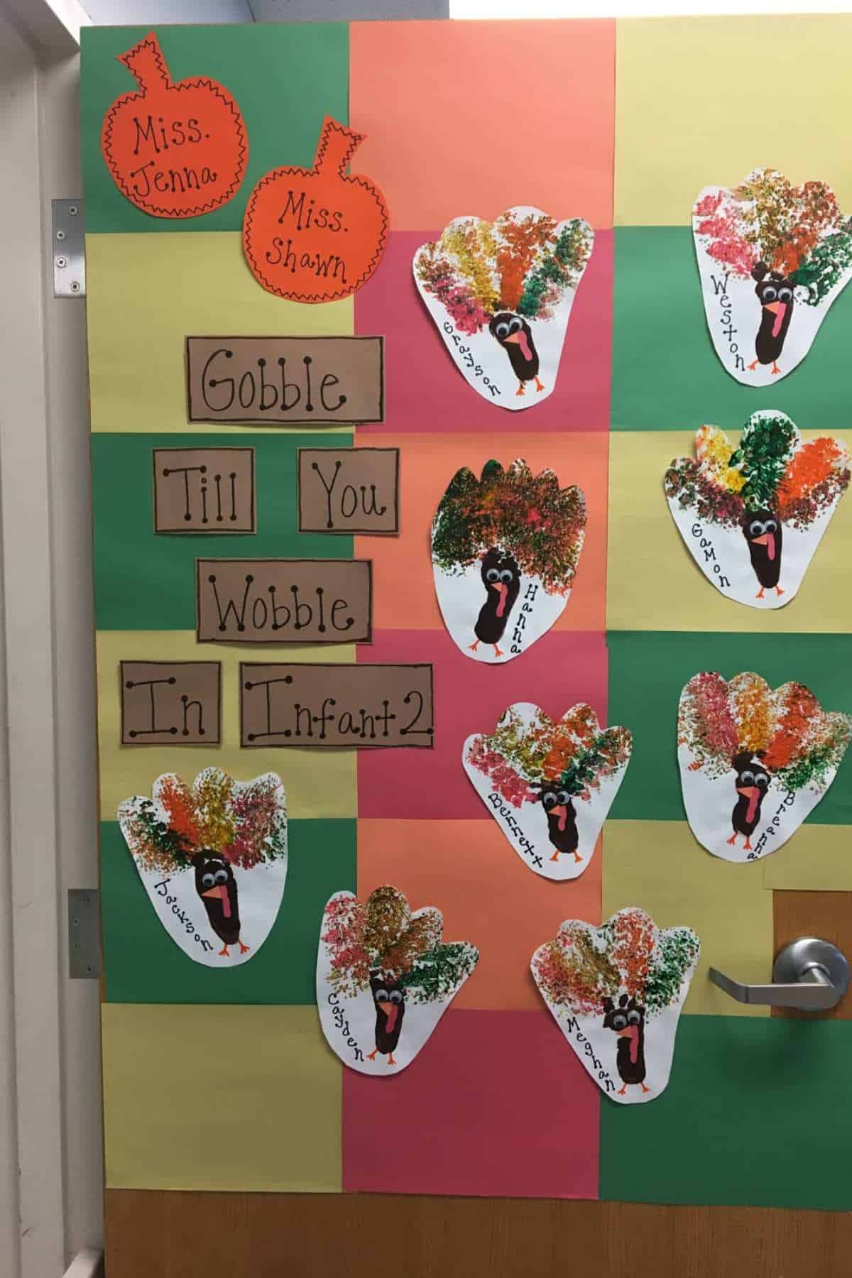 A door decorated with turkey crafts made from handprints, labeled "Gobble Till You Wobble in Infant 2," with teachers' names, Miss Jenna and Miss Shawn, on paper pumpkins.