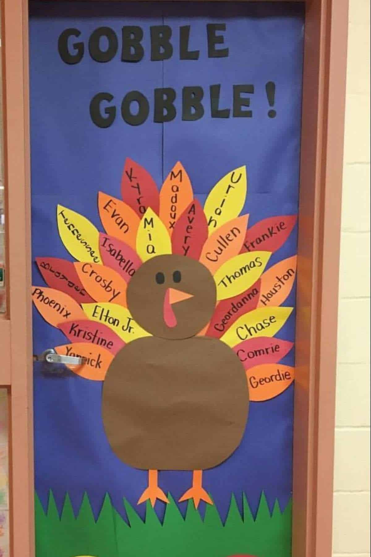 A classroom door decorated with a large paper turkey, its feathers labeled with student names, and the words "GOBBLE GOBBLE!" displayed at the top.