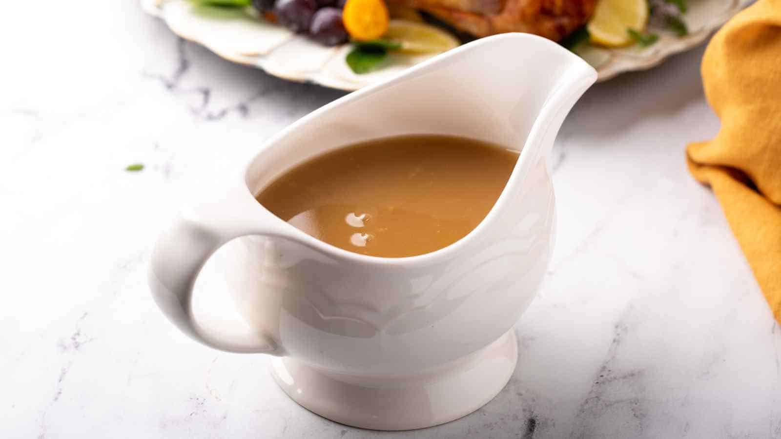 A white gravy boat filled with brown gravy sits on a marble surface, with part of a plate of food visible in the background.