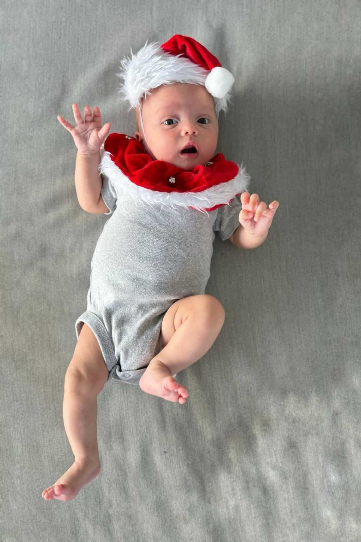 A baby lies on a grey surface wearing a light grey onesie and a red Santa hat with a matching collar, looking up with one hand raised.