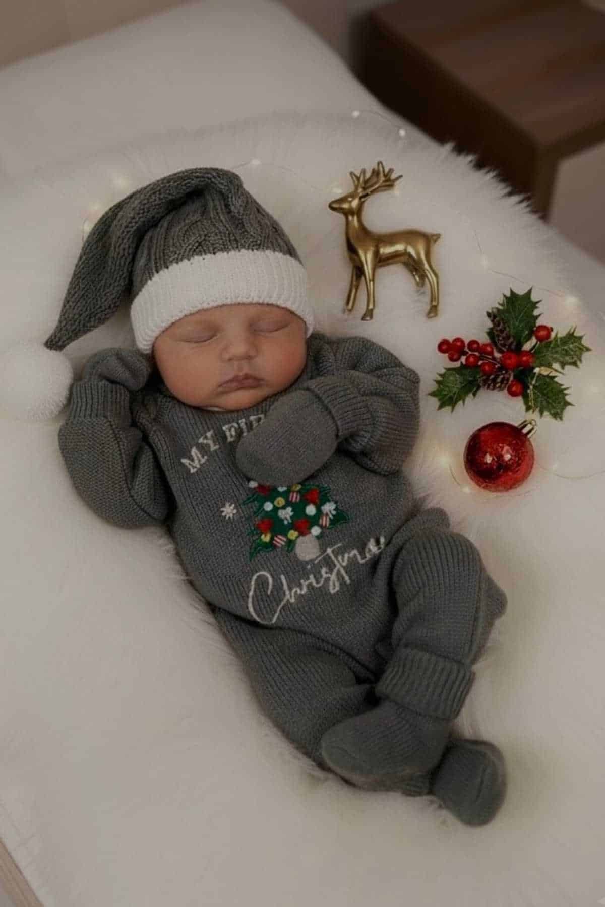 A baby in a gray knit Christmas outfit and hat sleeps on a white blanket beside a gold reindeer, holly, and red ornaments.
