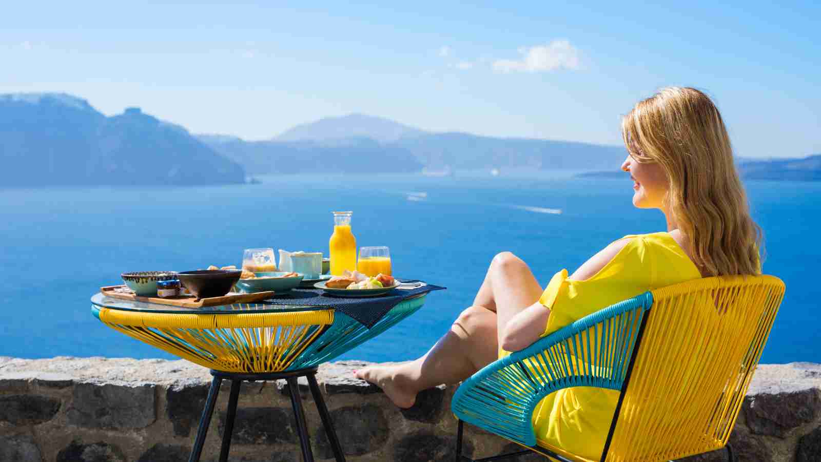 A woman in a yellow dress sits at a patio table with breakfast, overlooking the sea and distant mountains on a sunny day.
