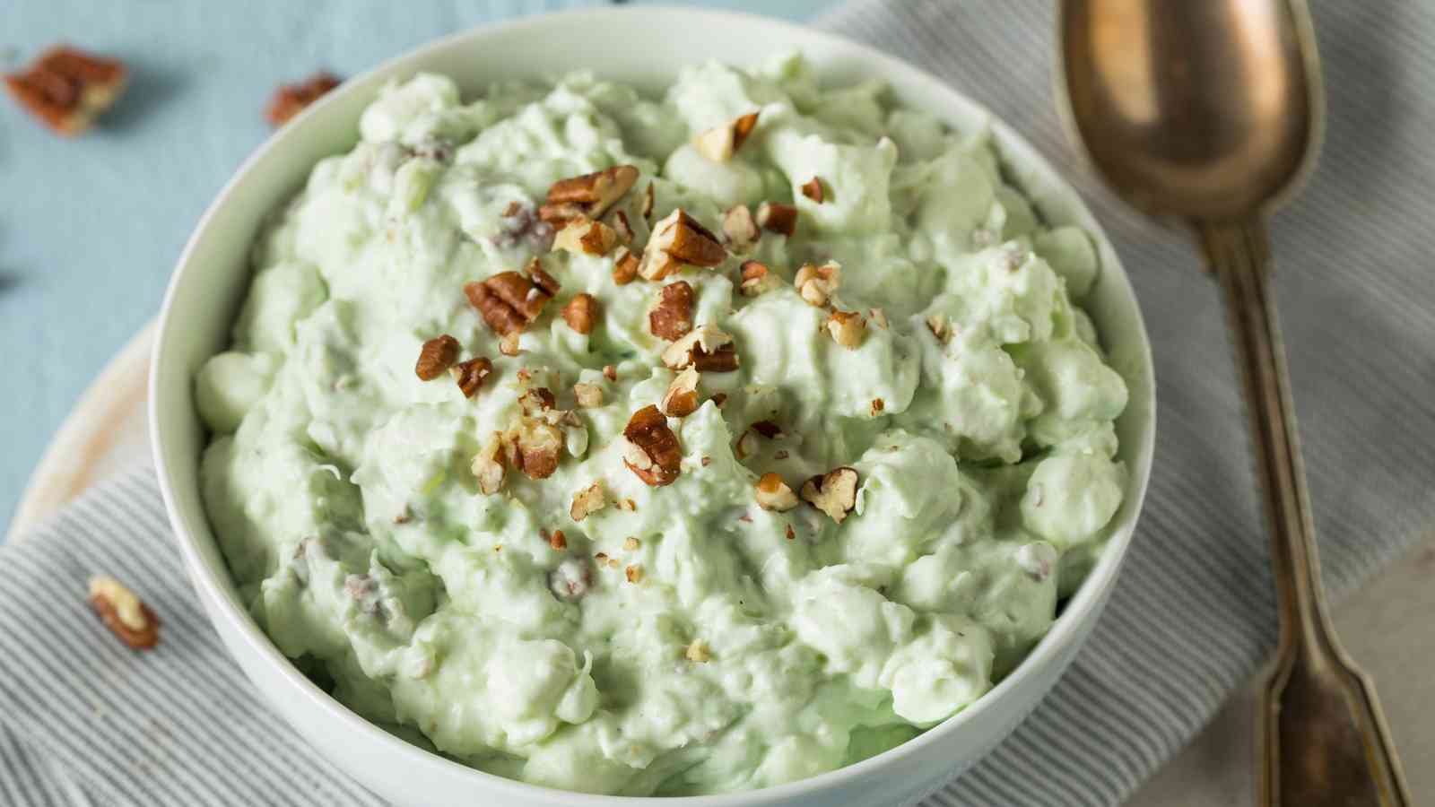 A white bowl of green pistachio salad topped with chopped nuts, placed on a striped cloth next to a metallic spoon.