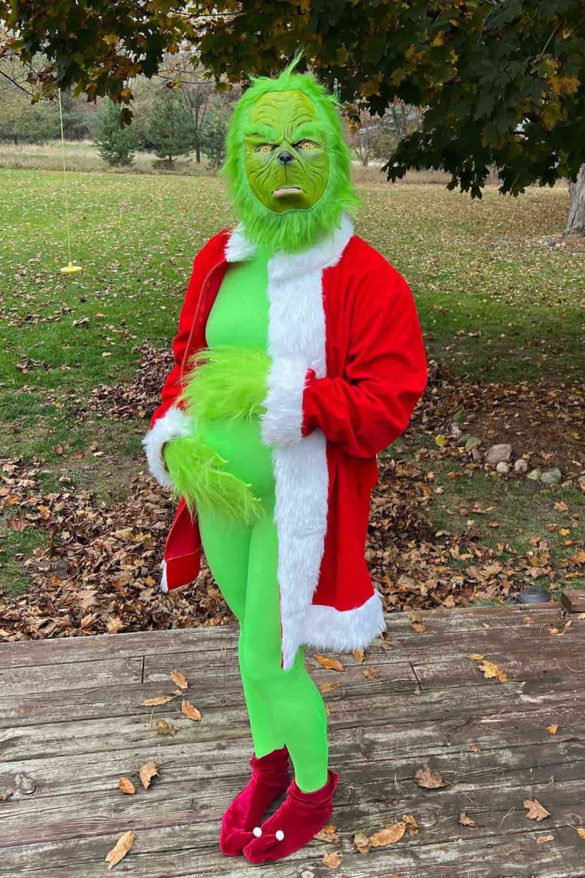 A person wearing a green Grinch costume, red Santa jacket, and red slippers stands on a wooden deck with autumn leaves on the ground behind them.
