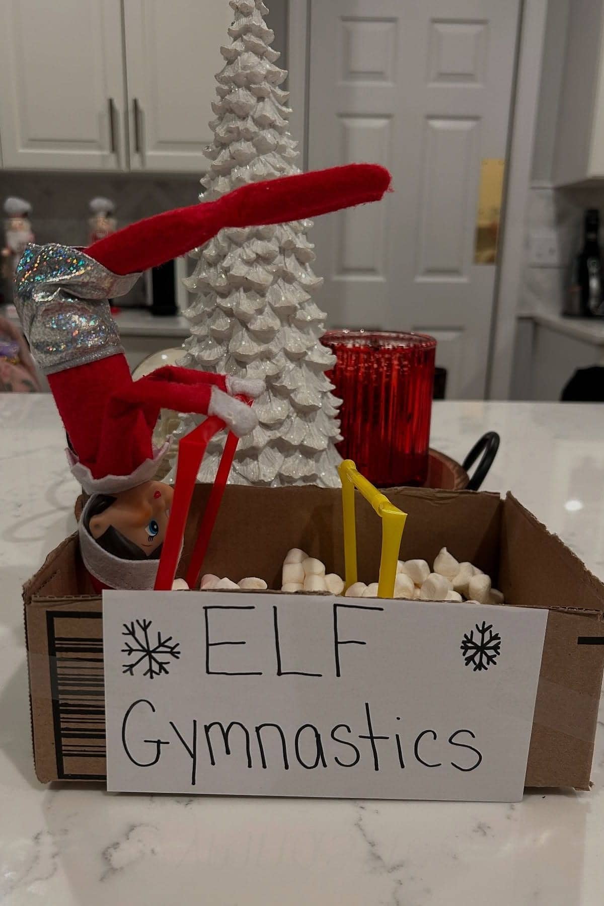 An Elf on the Shelf doll poses upside-down on homemade parallel bars made of straws in a box labeled "Elf Gymnastics," with marshmallows and a white Christmas tree in the background.