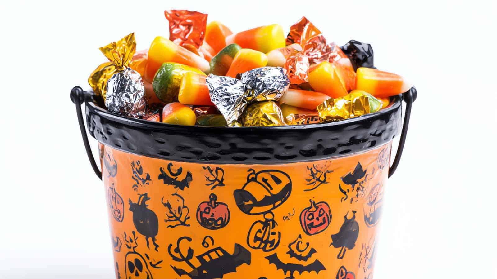 A metal bucket decorated with Halloween-themed images is filled with assorted wrapped candies and candy corn.