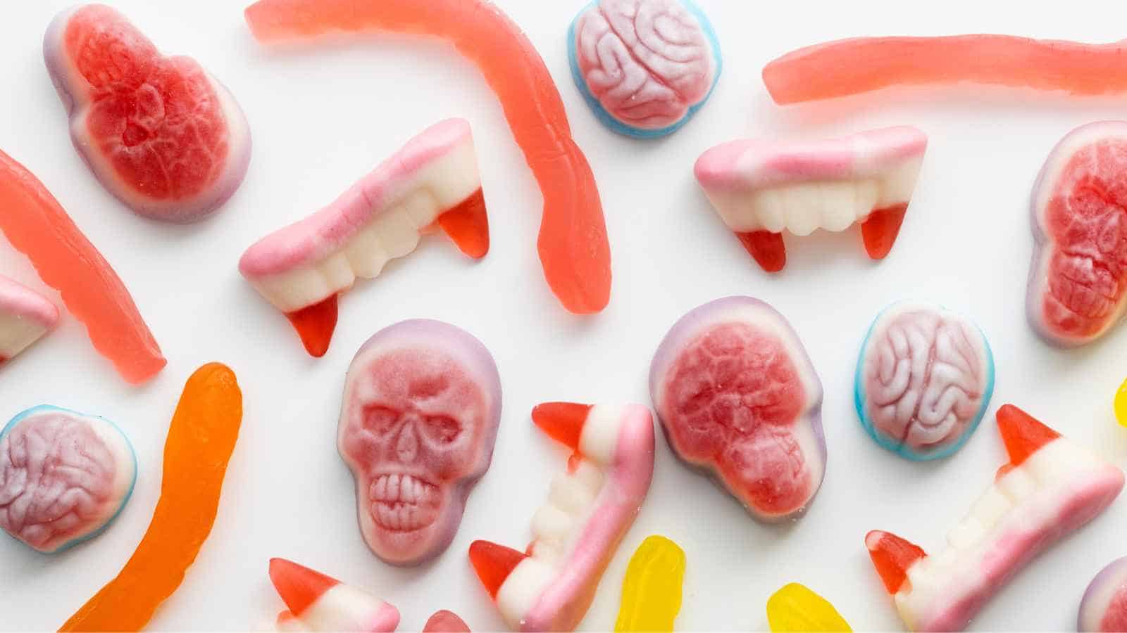 An assortment of gummy candies shaped like skulls, brains, fangs with dripping "blood," and worms, arranged on a white background.