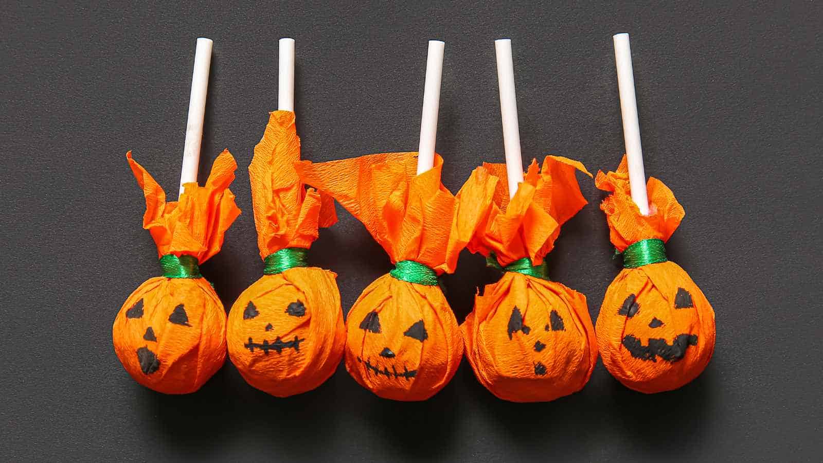 Five lollipops wrapped in orange paper with jack-o'-lantern faces and green ribbons, arranged in a row on a dark background.