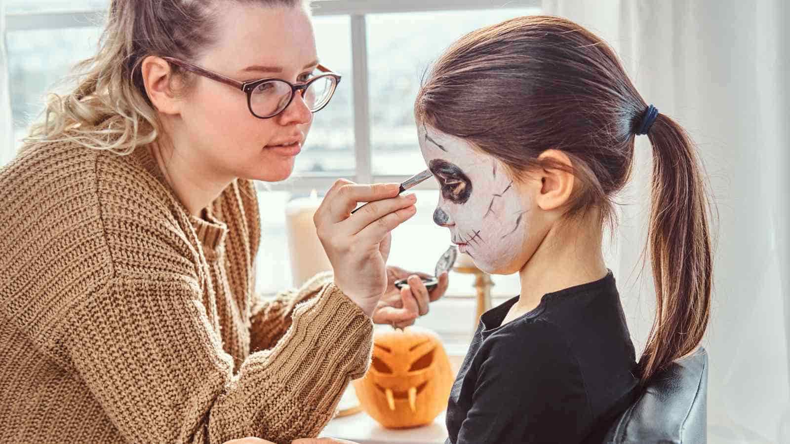 An adult paints a child's face with a skeleton design while a carved pumpkin sits on the table in the background.