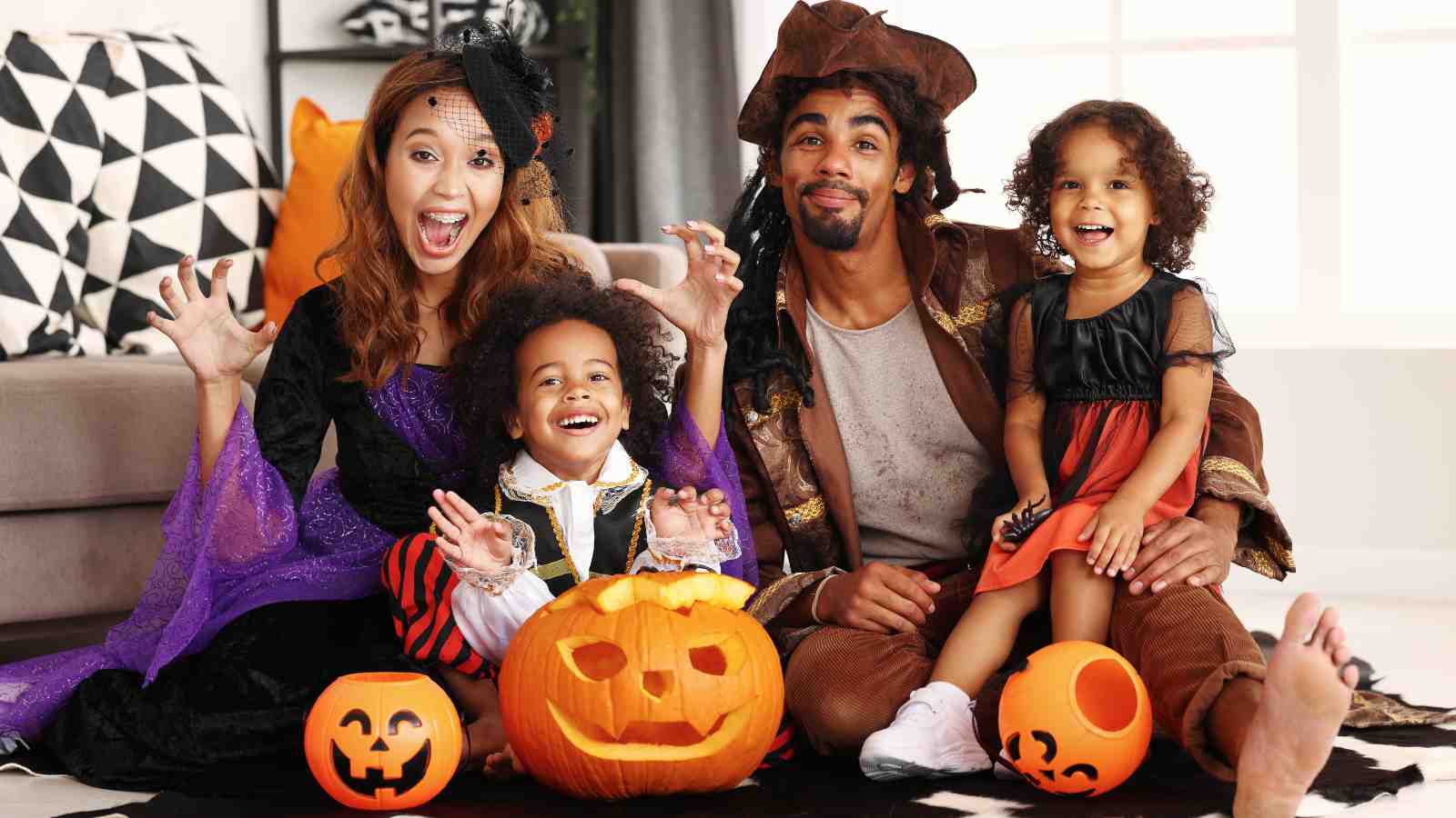 A family of four dressed in Halloween costumes sits on the floor with carved pumpkins, smiling and posing for the camera.