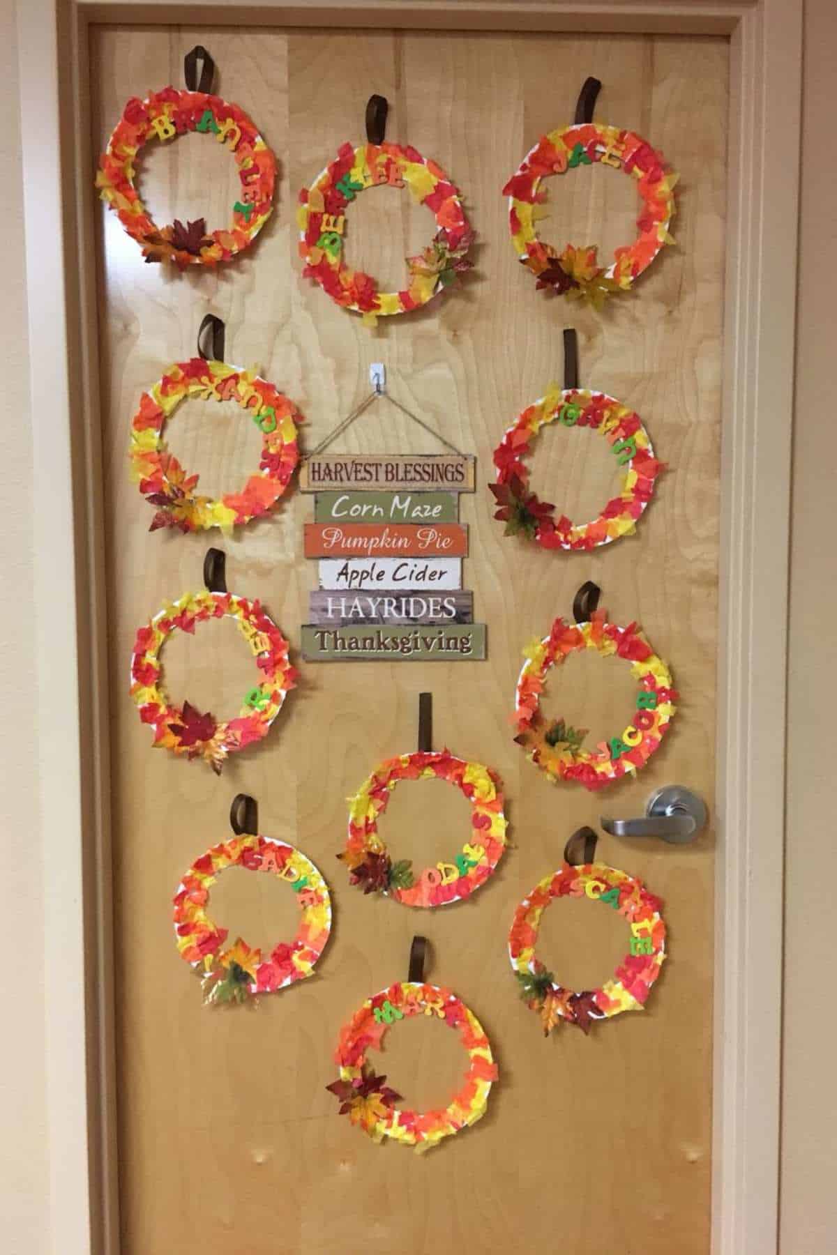 A wooden door decorated with paper wreaths made of colorful autumn leaves and a sign listing fall activities such as "Corn Maze," "Pumpkin Pie," "Apple Cider," "Hayrides," and "Thanksgiving.