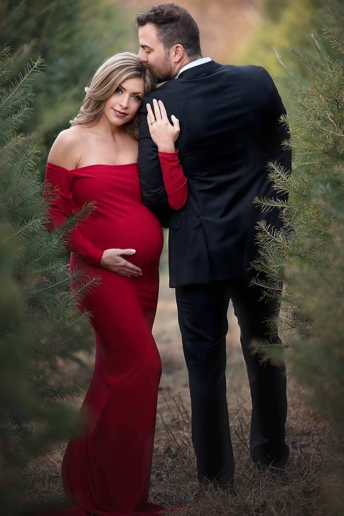 A pregnant woman in a red dress stands beside a man in a black suit, outdoors among green trees. The man embraces her and kisses her head.