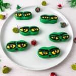 A white plate with six nutter butter cookies decorated with yellow and black eyes, surrounded by Christmas-themed candies and green tinsel on a white surface.