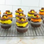 Chocolate cupcakes with orange and yellow frosting, each topped with a peanut butter cup, chocolate cookie, and candy, arranged on a cooling rack.