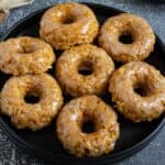 Eight glazed cruller-style donuts are arranged on a black plate set on a dark textured surface.