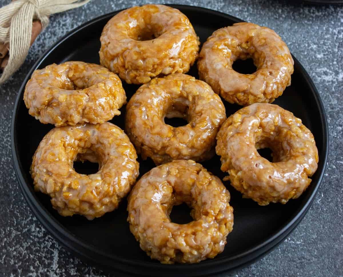 Eight glazed cruller-style donuts are arranged on a black plate set on a dark textured surface.