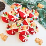A pile of red, Santa-themed chocolate-covered Oreos decorated with white icing, candy bears, and colorful candies, next to a green holiday-patterned cloth and plain Oreo cookies.