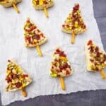 Tree-shaped white confections decorated with nuts and dried fruit, each with a pretzel stick as a trunk, arranged on parchment paper.