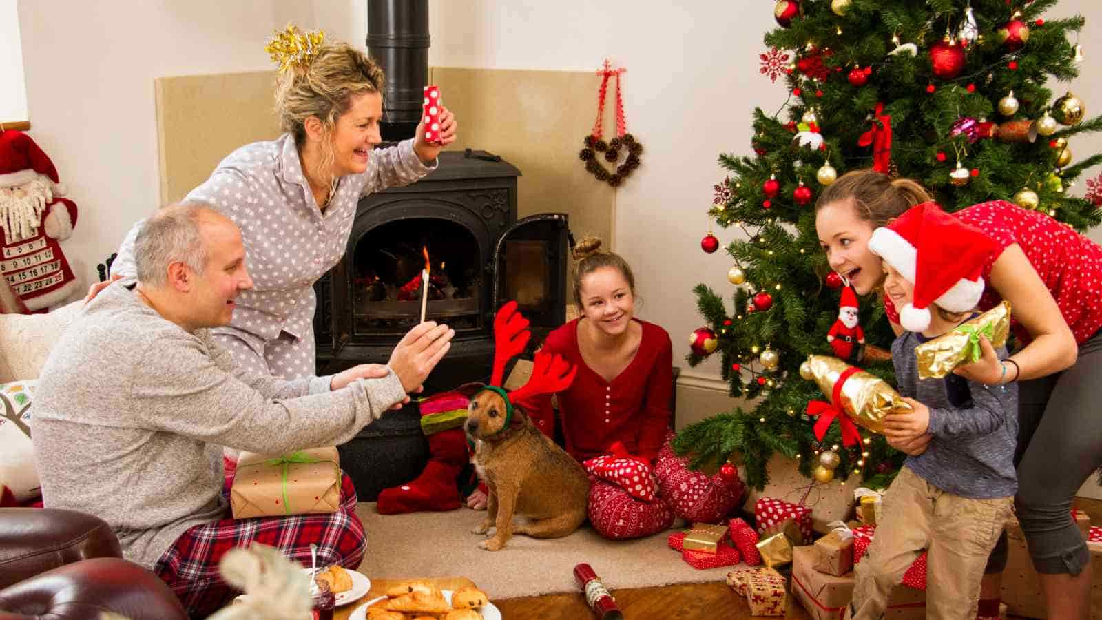 Four people and a dog sit by a fireplace and decorated Christmas tree, exchanging gifts and smiling, with holiday decorations and wrapped presents around them.