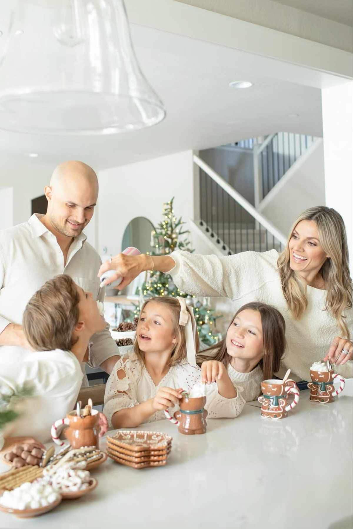 A family of five, all dressed in white, sits at a kitchen counter with hot chocolate and snacks, smiling and interacting in a holiday-decorated home.