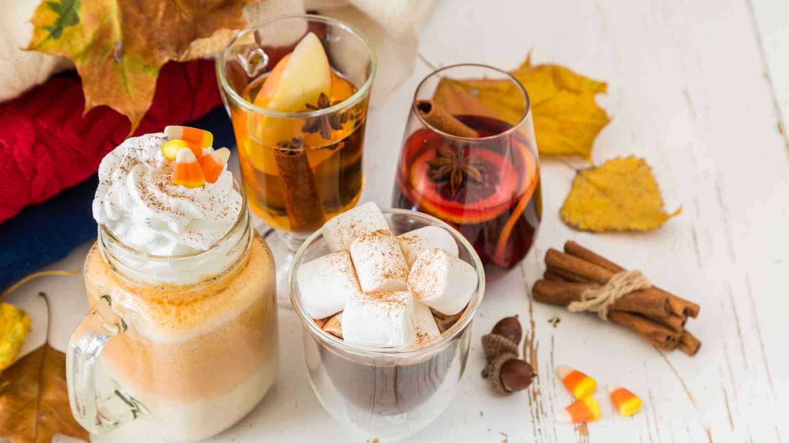 Four autumn-themed drinks, including a whipped-cream-topped beverage, hot chocolate with marshmallows, apple cider, and mulled wine, are arranged on a white table with fall leaves and spices.