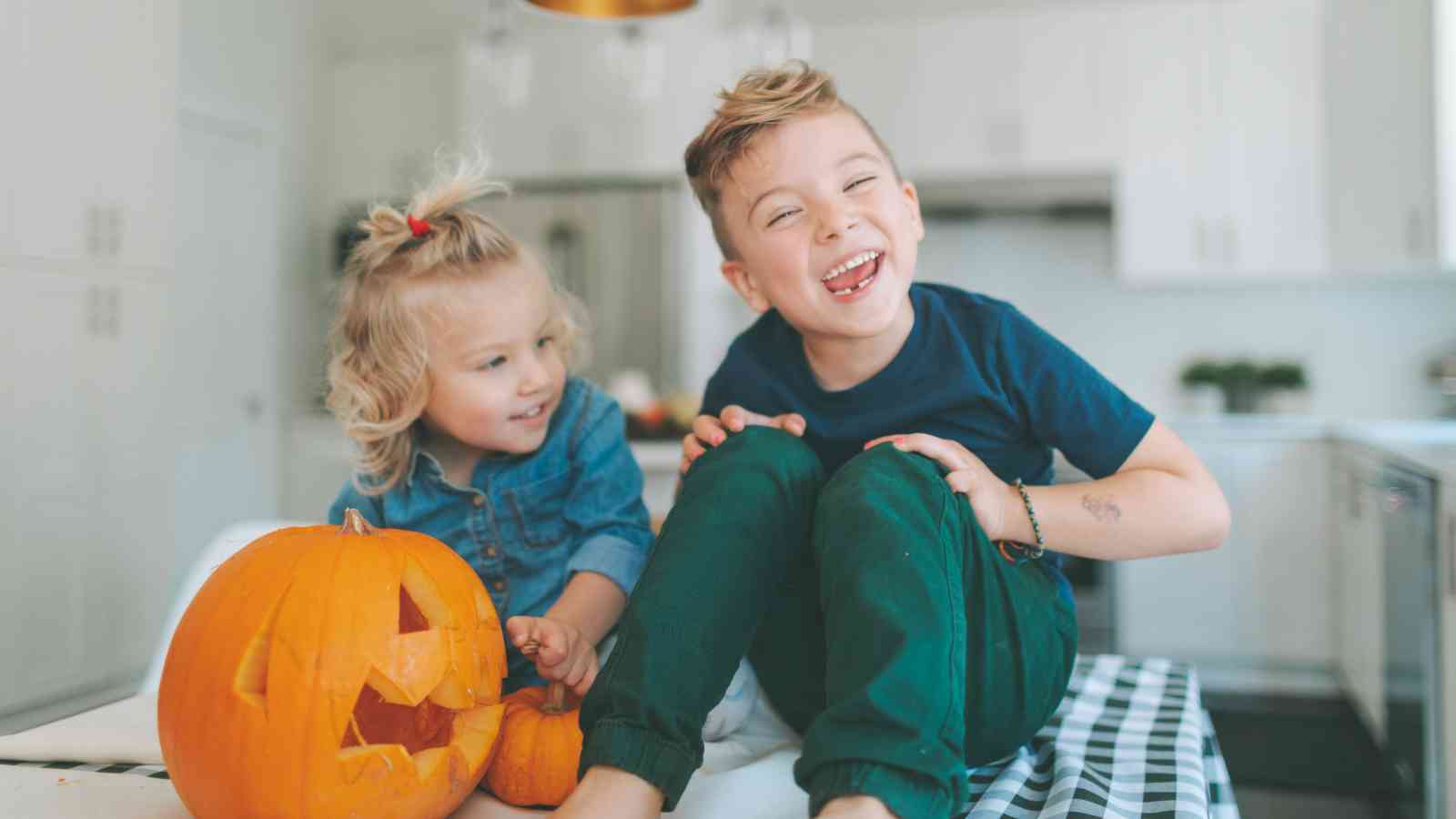 Two young children sit at a kitchen table with a carved jack-o'-lantern, smiling and laughing together.