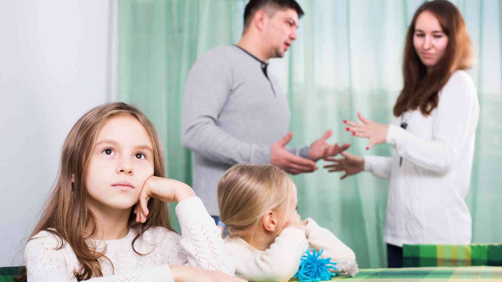 Two children sit at a table, one looking thoughtful and the other facing away, while two adults in the background appear to argue.