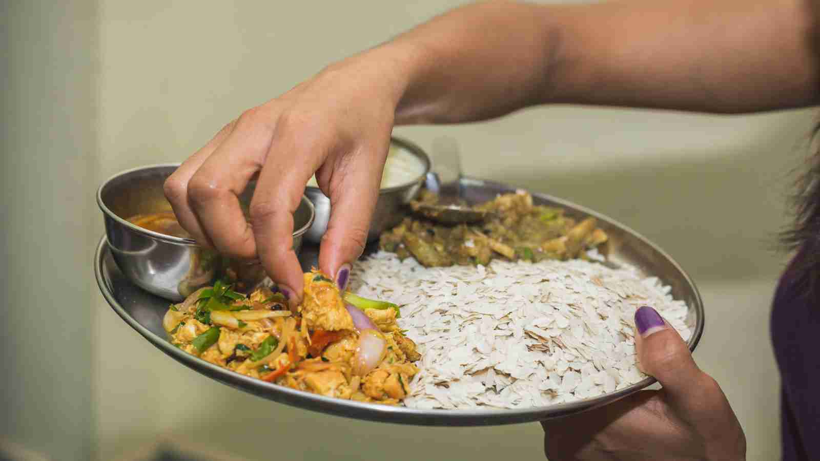 A person eating a traditional Indian meal with flattened rice, vegetables, dal, and curry served on a metal plate, using their hand to pick up food.