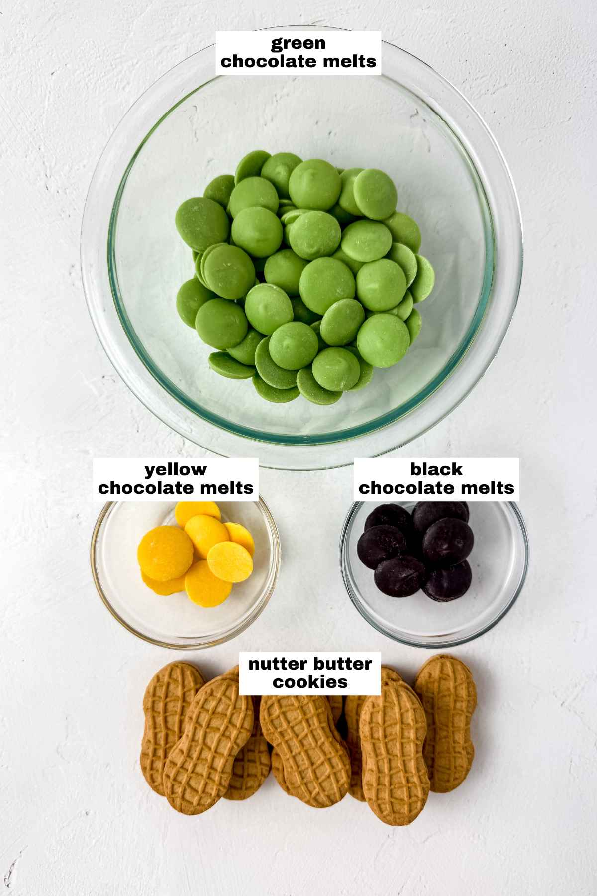 A glass bowl of green chocolate melts, a small bowl of yellow chocolate melts, a small bowl of black chocolate melts, and six Nutter Butter cookies on a white surface.