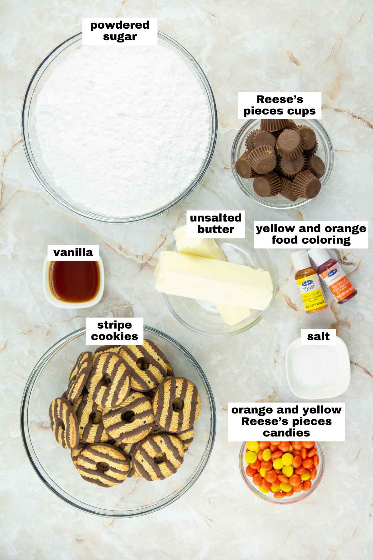 Various ingredients in bowls on a marble surface, including powdered sugar, stripe cookies, unsalted butter, vanilla, salt, Reese’s pieces cups, Reese’s pieces candies, and food coloring.