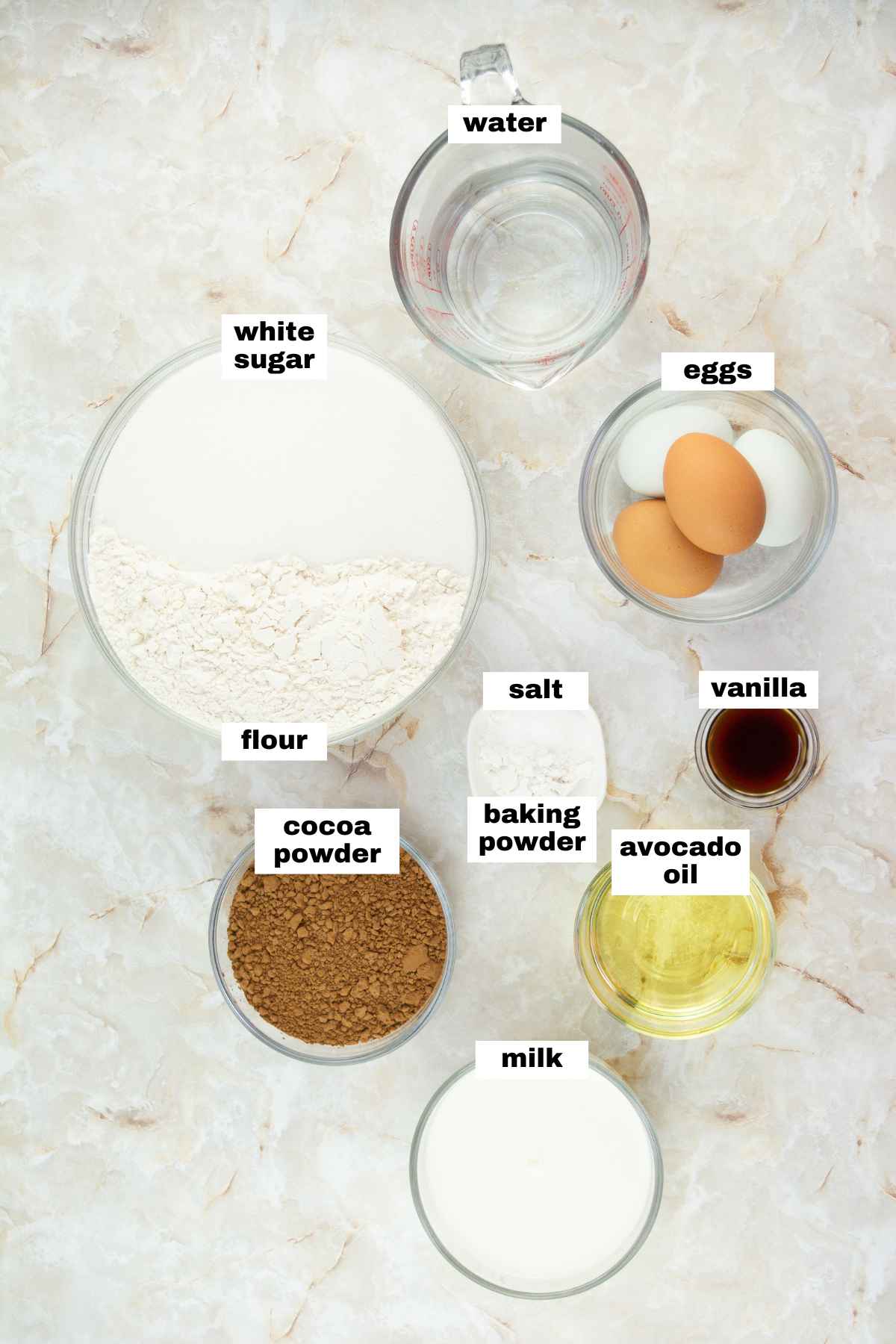 Top-down view of labeled baking ingredients in bowls and cups: white sugar, flour, cocoa powder, milk, water, eggs, salt, baking powder, vanilla, and avocado oil on a marble surface.