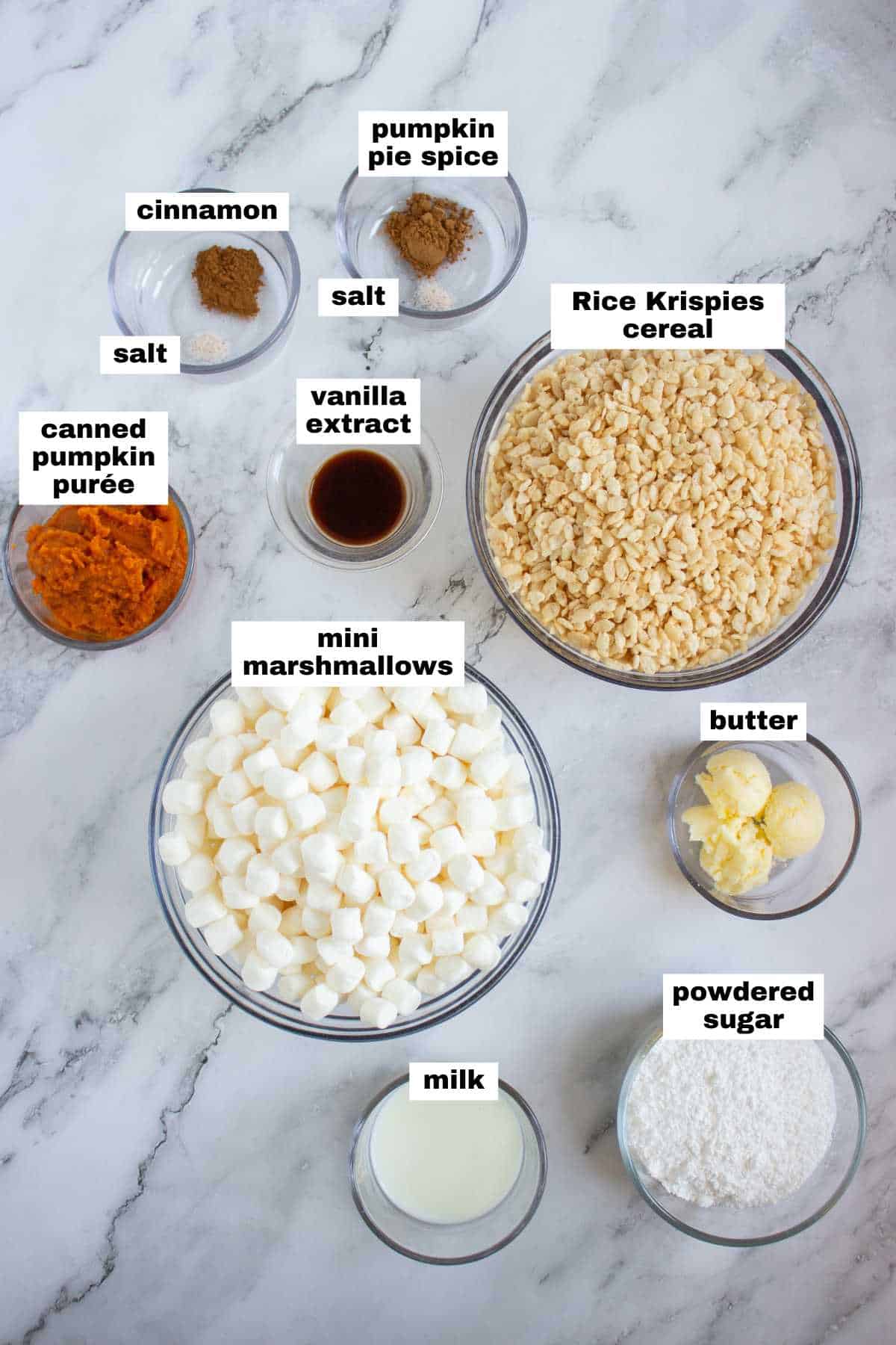Various ingredients for a recipe are arranged on a marble surface, including Rice Krispies cereal, mini marshmallows, canned pumpkin purée, butter, milk, powdered sugar, spices, and vanilla extract.