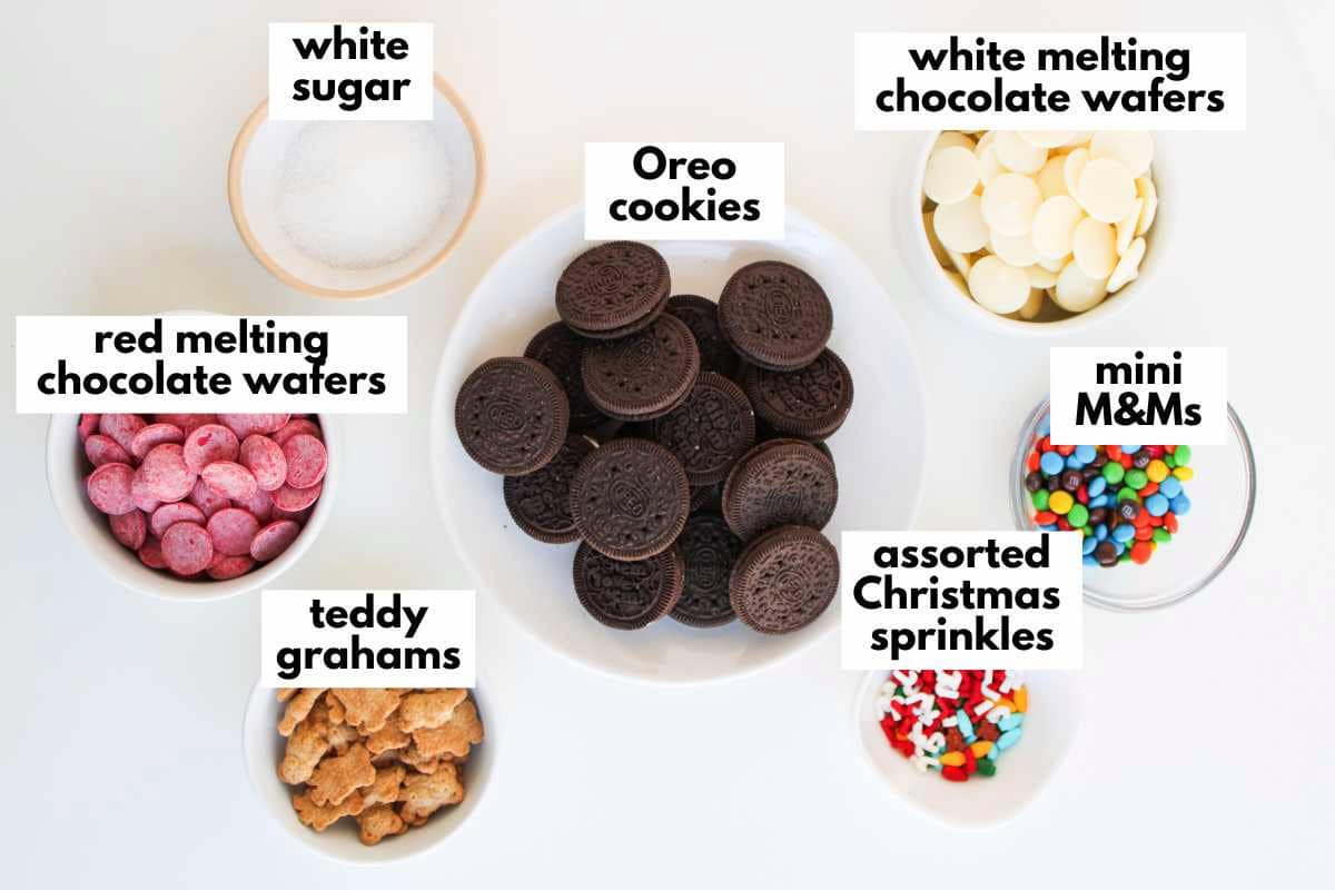 Bowls containing white sugar, Oreo cookies, white and red melting chocolate wafers, mini M&Ms, teddy grahams, and assorted Christmas sprinkles, each labeled.
