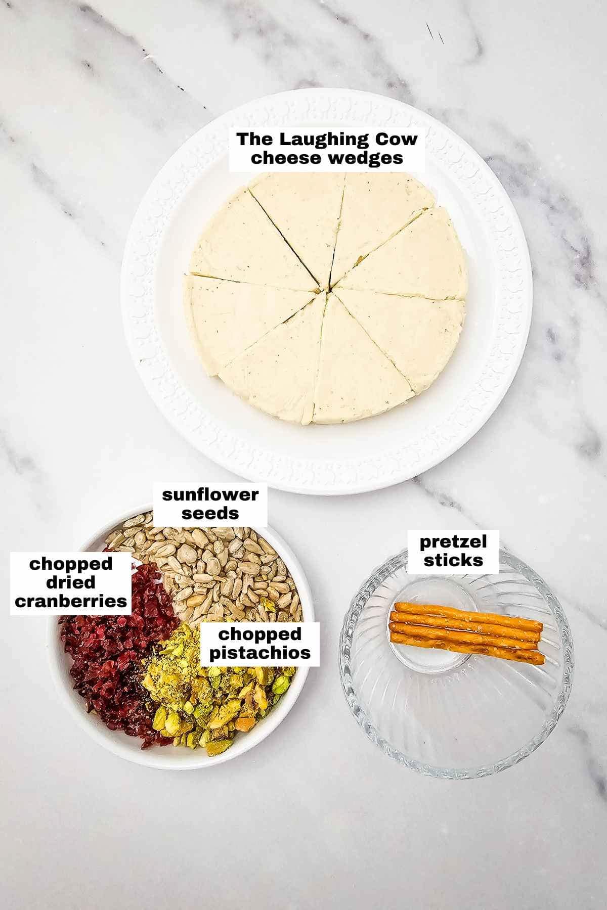 Three bowls on a marble surface: one with wedge-sliced cheese, one with sunflower seeds, cranberries, and pistachios, and one with pretzel sticks. Labels identify each item.