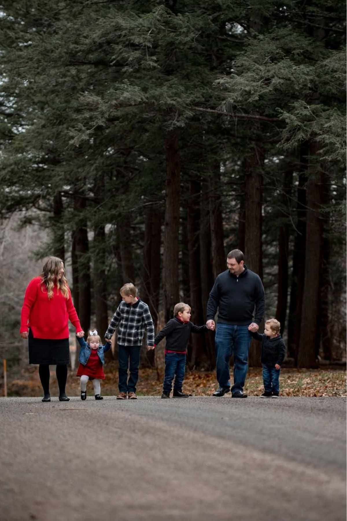 A family of six, including two adults and four children, walks hand in hand on a road through a wooded area.