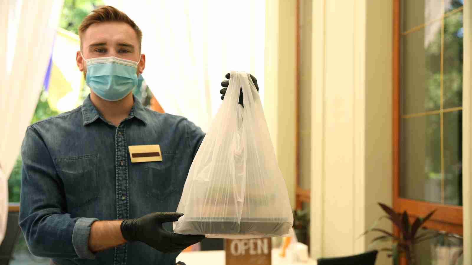 A person wearing a face mask and gloves holds out a plastic bag with takeout food containers inside, standing indoors near a window.