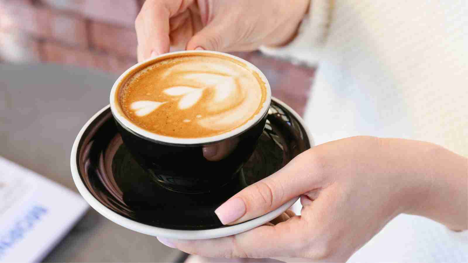 A person holding a black cup of latte with foam art in the shape of hearts, on a matching black saucer.