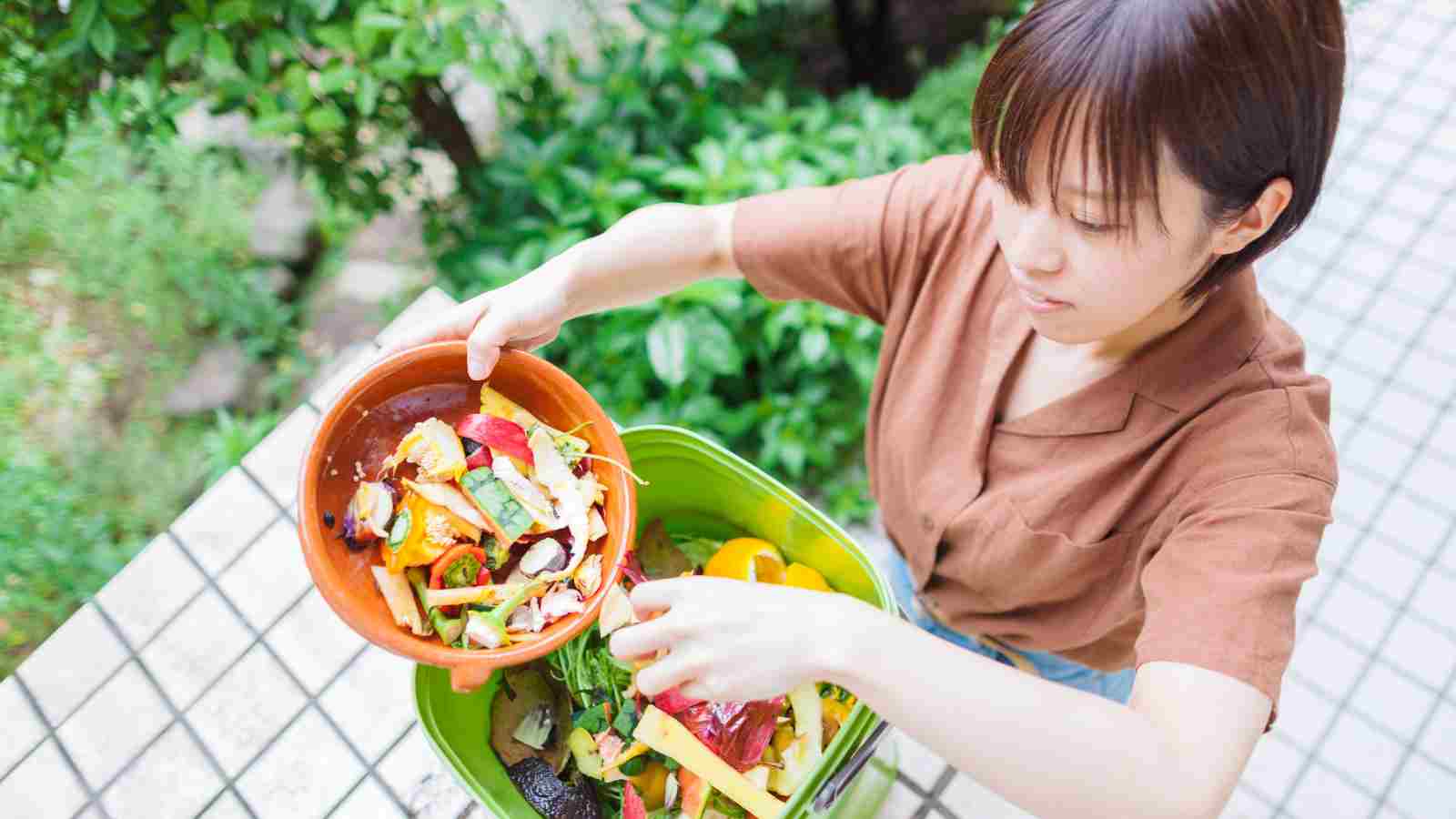 A person empties a bowl of food scraps into a green outdoor compost bin, surrounded by greenery and tiled ground, reflecting practices seen in countries that handle food waste better than the US.