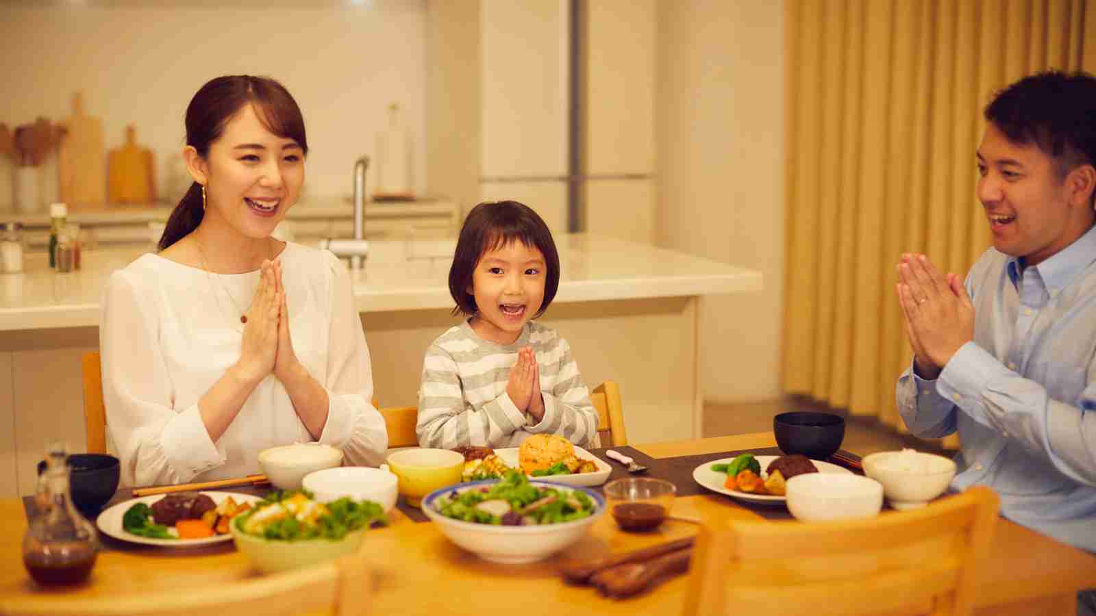 Three people sit at a dining table with various dishes, smiling and holding their hands together in a gesture before eating.