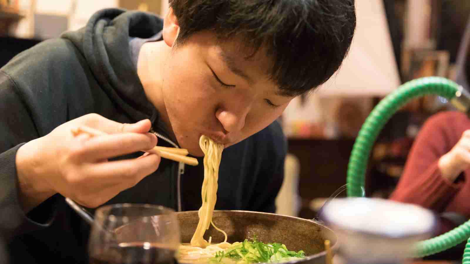 A person eats noodles from a bowl using chopsticks, leaning forward and slurping the noodles.