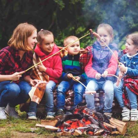 A woman and four children sit by a campfire roasting marshmallows, with a tent nearby and trees in the background.