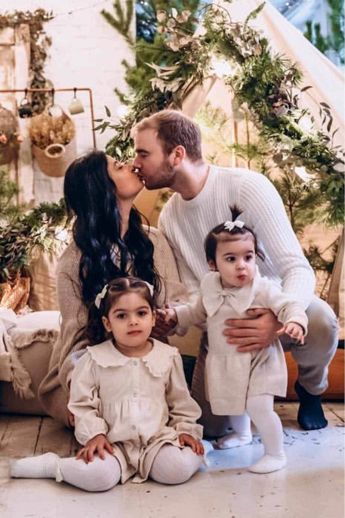 A man and woman kiss while kneeling behind two young girls dressed in matching light outfits, with greenery and holiday decorations in the background.