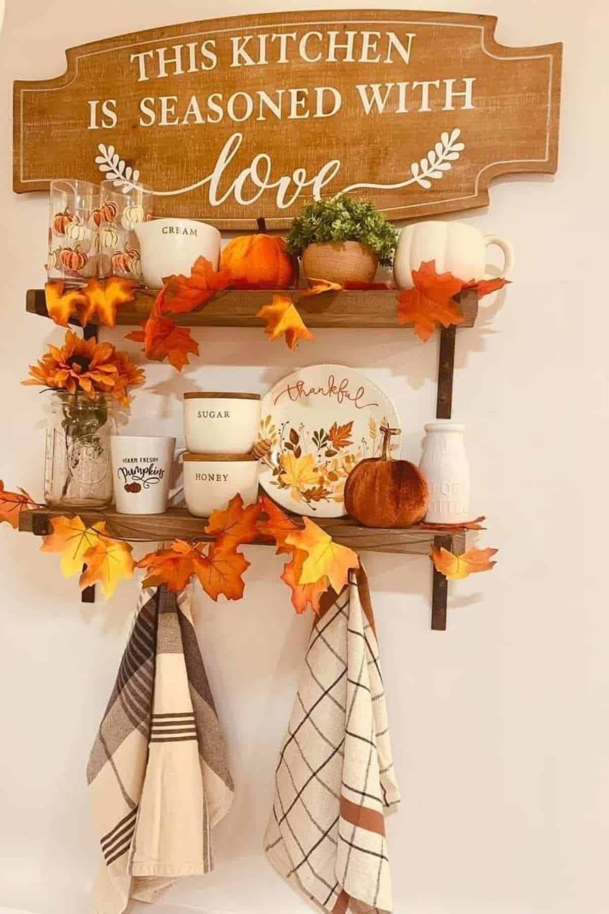 Two wooden shelves with mugs, jars, and fall decorations beneath a sign reading "This kitchen is seasoned with love"; dish towels hang below.