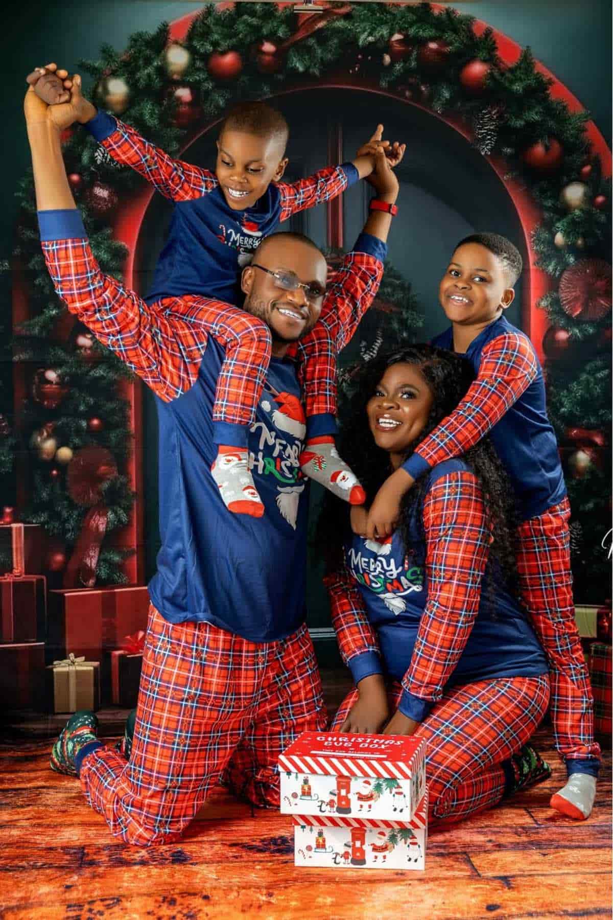 A family of four in matching plaid pajamas poses together in front of a Christmas backdrop, with wrapped gifts and festive decorations in the background.