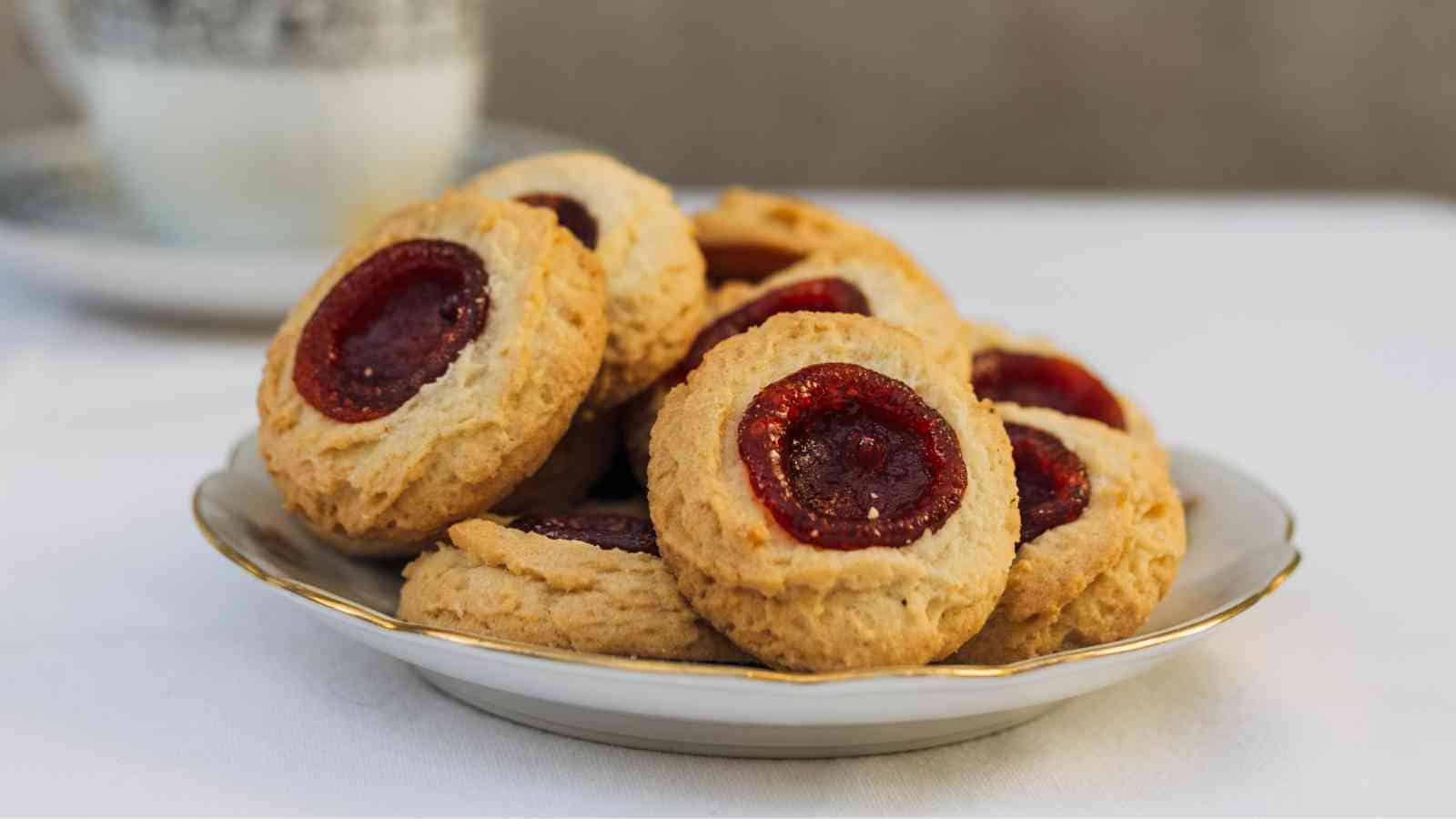 A white plate with several round cookies, each topped with a dollop of red jam, sits on a white tablecloth. A blurred cup and saucer are in the background.