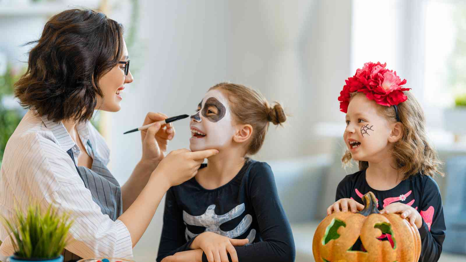 An adult paints a skeleton face on a smiling child, while another child with a painted spider on her cheek and a flower headband sits nearby with a carved pumpkin.